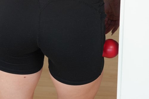 Woman using a red massage ball against a wall to target the glute muscles. This self-massage technique helps release tension, improve mobility, and reduce muscle soreness in the hips and lower back.