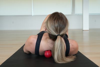 Woman lying on a yoga mat using a red massage ball to apply pressure to her upper back. This self-massage technique helps release tension, relieve muscle soreness, and improve mobility in the shoulders and spine.
