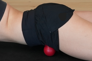 Woman lying face down on a mat using a red massage ball to apply pressure to her hip flexor muscles. This self-massage technique helps release tension, improve mobility, and reduce muscle tightness in the hip area.