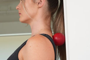 Woman using a red massage ball against a wall to apply pressure to her upper back and neck. This self-massage technique helps release tension, improve posture, and relieve muscle tightness.