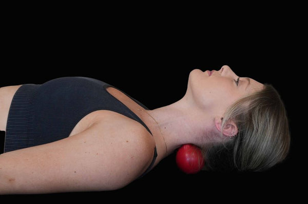 Woman lying down using a red massage ball for neck tension relief. The ball is positioned under the base of her head, providing support and pressure to alleviate neck pain. Black background emphasizes the therapeutic focus. Ideal for promoting neck massage techniques and self-care practices.